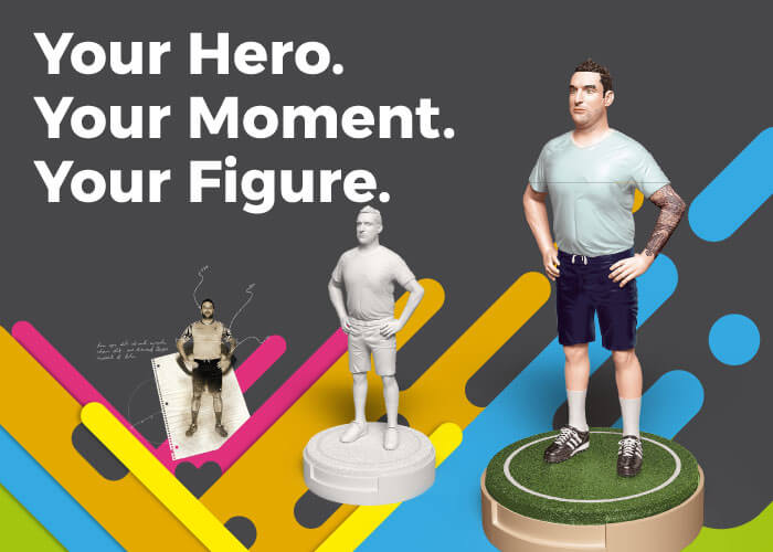 Helden-Figur als Highlight: Bunter dynamischer Hintergrund mit Illustration, Weißmuster und fertig bemalter Hero-Figur. Slogan ‚Your Hero. Your Moment. Your Figur.‘ auf der Messewand